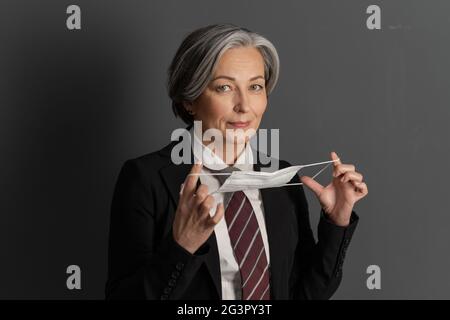 Charming gray-haired business woman putting on protective mask. Mature Caucasian woman wearing suit with tie looking at camera. Stock Photo