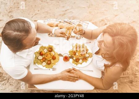 Loving mature couple have romantic date in cafe on sandy beach outdoor. Man and Woman holding hands sit at table with delicious Stock Photo