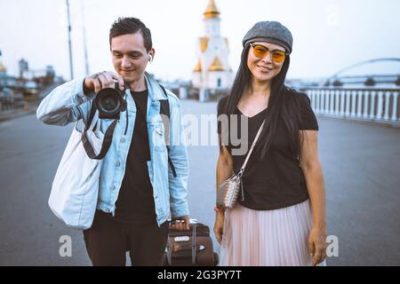 Happy tourists with suitcase on embankment of river. Young man takes photo. Woman wearing casual clothes smiling looking at came Stock Photo