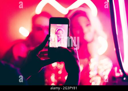 Young woman is photographed in a night club. Focus on male hands making photo by smartphone in foreground. Abstract blurred back Stock Photo