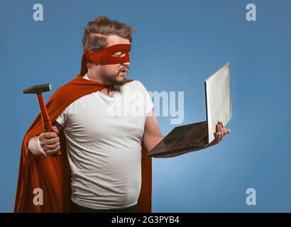 Super hero man going to smash or repair laptop computer with hammer. Side view of masked man in red super hero uniform posing on Stock Photo