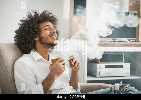 Handsome man enjoys relaxing holding cup of drink in his hands. Young guy exhales cloud of smoke smoking an electronic cigarette Stock Photo