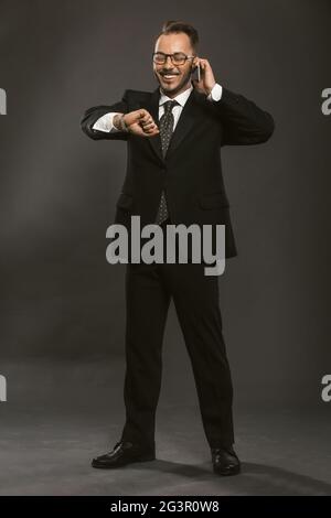 Happy businessman smiles checking time on wristwatch while talking on mobile phone. Caucasian handsome looks at hand clock waiti Stock Photo