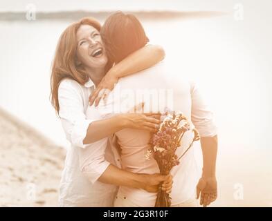 Happy couple hugs on the seashore. Laughing woman hugs a man holding a bouquet of flowers behind him on a summer evening. Surpri Stock Photo