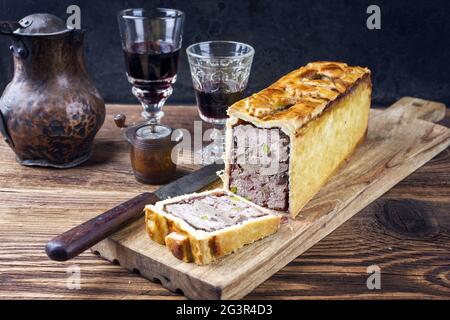 Traditional French Pate en croute with goose meat and liver as closeup with red wine on a wooden board Stock Photo