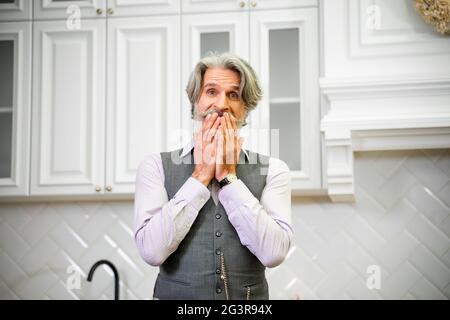 Elegant senior grey haired grandfather in formal wear being surprised when receiving present for his birthday, standing in stylish kitchen at home holding hands over mouth with excited expression Stock Photo