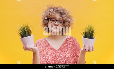 Attractive girl holding two small pot plants in her hands. Girl wearing round spectacles dressed in casual pink top with flowers in her curly hair. Isolated with yellow background studio. Stock Photo