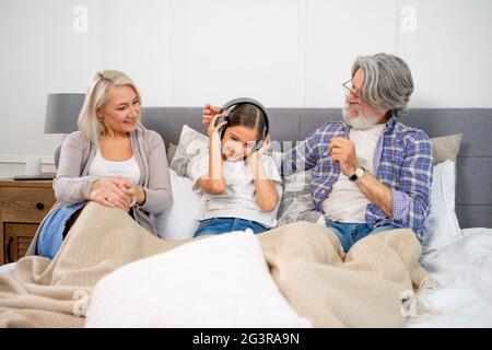 Cute little girl granddaughter listening music in headphones with her positive grandparents while lying on bed, having fun and relaxing together. Happy family enjoying leisure time on weekend at home Stock Photo