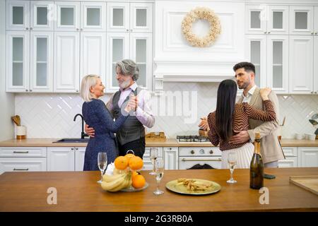Two couples grandparents and young husband and wife dancing in light stylish kitchen near table with champagne during birthday celebration, having fun and enjoying time together at home party Stock Photo