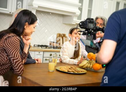 Big multigenerational family having photo or video shoot when cute smiling little girl daughter posing for camera sitting at kitchen table with grandparents and mother. Birthday celebration concept Stock Photo