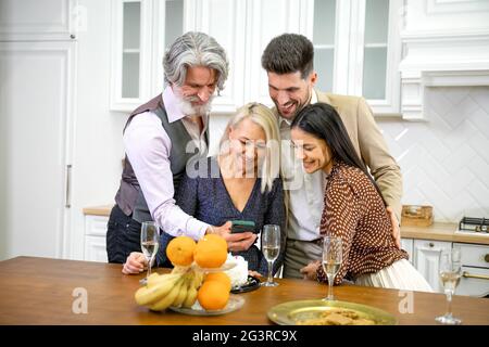 Positive grandparents and smiling young couple in formal wear looking at pictures and videos on smartphone during birthday celebration with family at home in kitchen with champagne on table Stock Photo