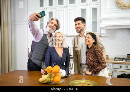 Happy family grandparents and young smiling couple in formal festive wear taking selfie on smartphone, looking at camera and smiling during birthday celebration in kitchen at home Stock Photo