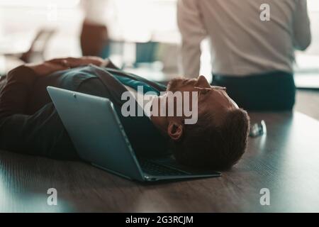 Tired office worker is lying on his desktop on his laptop. He's wearing glasses and a tie suit. In the background his colleague Stock Photo