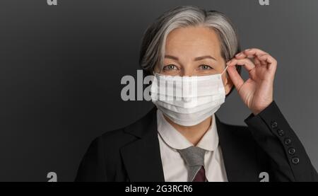 Pretty gray-haired middle-aged business woman putting on protective mask and showing ok or okey sign while posing on gray wall b