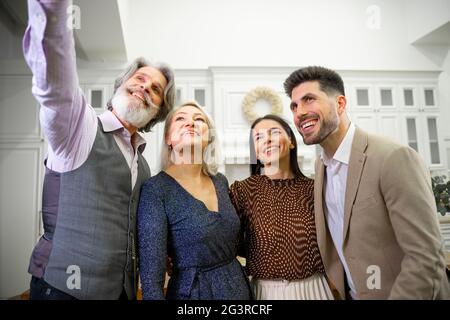 Happy family, smiling grandparents and young married couple taking selfie on grandfathers smartphone during birthday celebration in stylish kitchen at home, expressing positivity and happiness Stock Photo