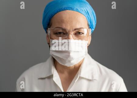 Female doctor face, close up shot. Mature woman in protective mask and goggles looks at camera. Isolated on gray Stock Photo