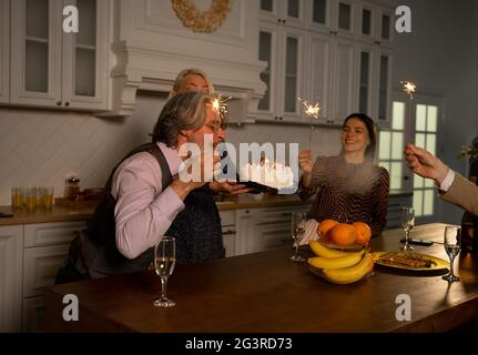 Grandfather blowing out candles on birthday cake and making wish while sitting behind kitchen table at home with his wife holding cake and guests holding sparklers. Family celebration concept Stock Photo
