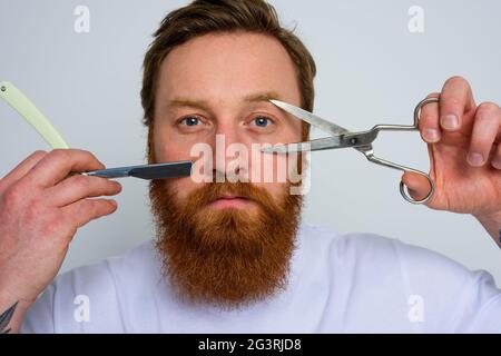 Worried man with scissors and blade is ready to cut the beard Stock Photo