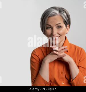 Mature woman in orange shirt smiles at camera. Female hands are folded into the castle. Isolated background of photo studio. Hig Stock Photo