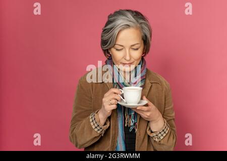Charming elegant woman drinks coffee or tea holding white cup looking on it. Isolated on pink background with copy space. Studio