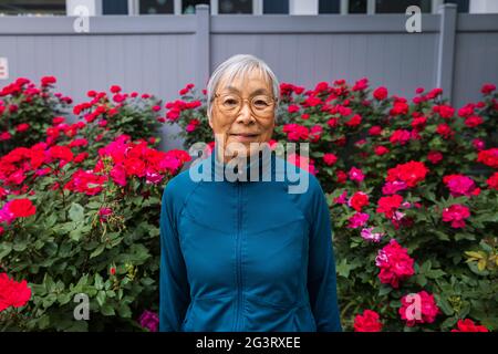 Senior woman wearing eyeglasses standing in front of red flower garden Stock Photo