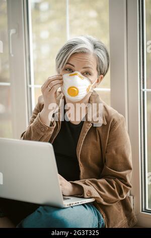Mature woman in protective mask works laptop sitting on sill Stock Photo