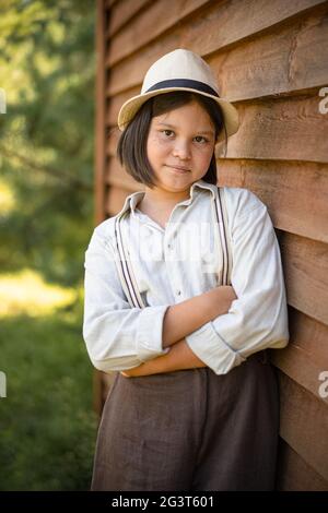 Country girl poses leaning against wooden plank wall of building. Teenage girl in white hat and braces looks at camera crossed a Stock Photo