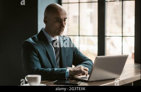 Bald middle aged businessman in business suite working on laptop next to open windows in modern office. Handsome bold man workin Stock Photo