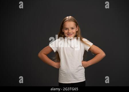 Bossy little girl of 8,10 years put her hands sideways looks questioningly at the camera wearing white t shirt isolated on dark Stock Photo