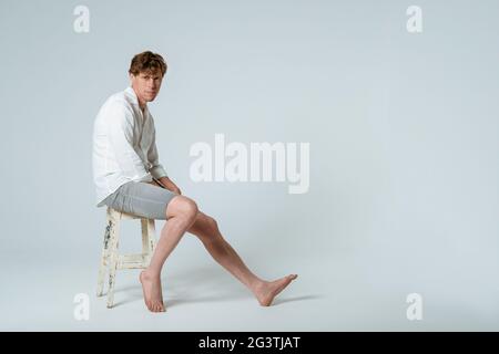 Full length shot of young handsome young man sitting on chair wearing white shirt and grey shorts with one leg out looking at ca Stock Photo