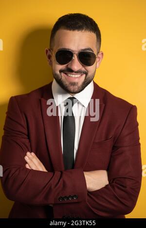 Close up portrait of handsome business man in sunglasses and burgundy suit looking at camera with hands folded isolated on yello Stock Photo