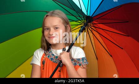 Little girl in orange dress holding rainbow colors umbrella standing cheering in the studio looking at the camera isolated on da Stock Photo