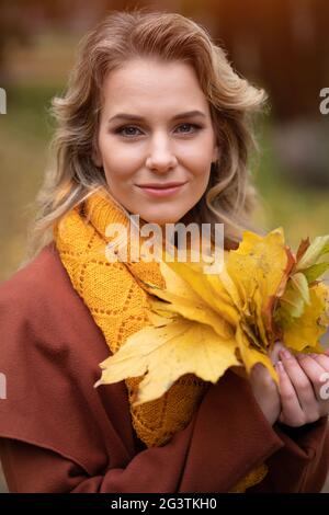 Beautiful young woman posing for camera with autumn leaves in hand and fall yellow garden or park. Beautiful smiling young woman Stock Photo