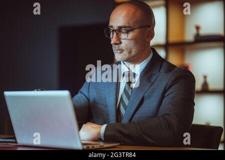 Confident bald middle aged CEO working on laptop wearing business suite sitting in modern office. Handsome bold man working on l Stock Photo