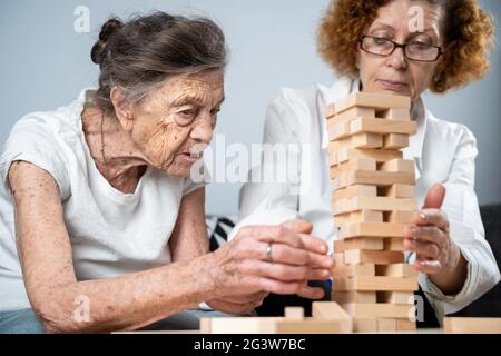 Senior woman playing Jenga, build tower of blocks. Elderly doctor in white lab coat, supporting senior patient, developing logic Stock Photo