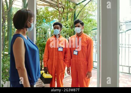 Mature Asian woman in mask opening door to multi-ethnic house servicemen in orange bodysuits Stock Photo