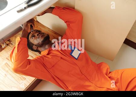 Concentrated young Black bearded plumber with badge on chest lying under sink and fixing clogged drains Stock Photo