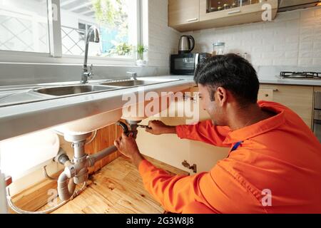 Busy young mixed race plumber in orange bodysuit unscrewing water pipe while changing it under sink in kitchen Stock Photo