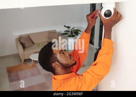 Serious young African American technician in orange wear using screwdriver while screwing security camera on ceiling Stock Photo