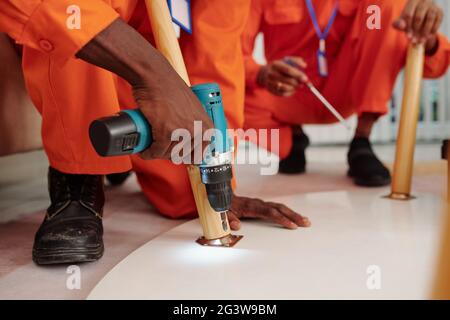 Close-up of unrecognizable Black handyman in orange wear installing screw into table using cordless drill while assembling table with colleague Stock Photo