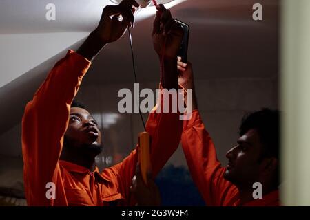 Afro-American electrician in orange suit measuring electrical power while inserting light bulb with coworker holding phone with flashlight Stock Photo