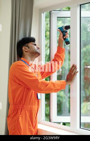 Busy young Indian window installation specialist in orange bodysuit using cordless drill while fitting window in flat Stock Photo