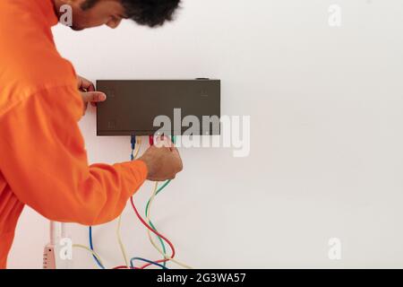 Over shoulder view of serviceman in orange wear plugging connector in transmitter box on wall while mounting it Stock Photo