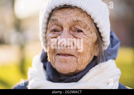 Portrait Caucasian senior woman with gray hair and deep wrinkles 90 years old posing in warm clothes, white knitted scarf and ha Stock Photo