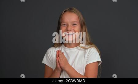 Happy charming little girl with hands put together looking a side of camera, wearing white t-shirt isolated on dark grey or blac Stock Photo