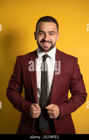 Young handsome man wearing amazing elegant burgundy suit looking on you smiling with hands holding a jacket isolated on yellow b