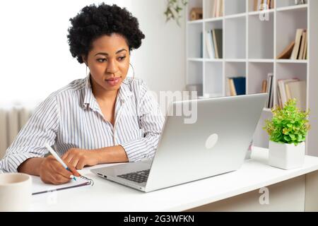African American female student uses a laptop and writes in a notebook. Portrait of a young black woman working remotely on the computer at home. Stock Photo