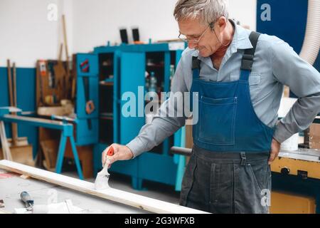 An elderly cabinetmaker in overalls and glasses paints a wooden board with a brush on a workbench in a carpentry shop. Stock Photo