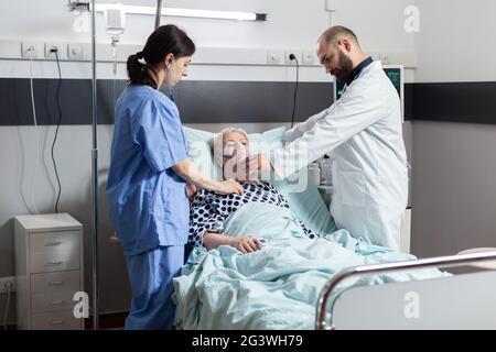 Hospitalized senior woman inhale and exhale through oxygen mask laying in hospital bed. Medical nurse using stethoscope listening patient heart. Eldely woman with oximeter attached on finger. Stock Photo