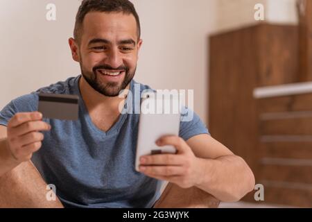 Using smartphone handsome bearded guy holding debit or credit card in hands buying online. Modern digital life. Digital app on s Stock Photo